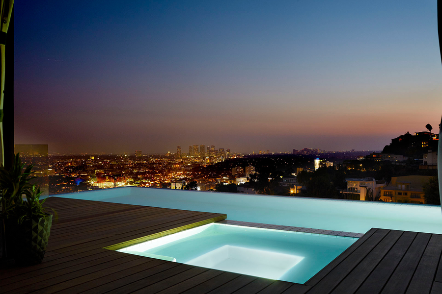 Modern rooftop infinity pool and hot tub overlooking a glowing city skyline at dusk, with soft ambient lighting enhancing the luxurious outdoor setting.